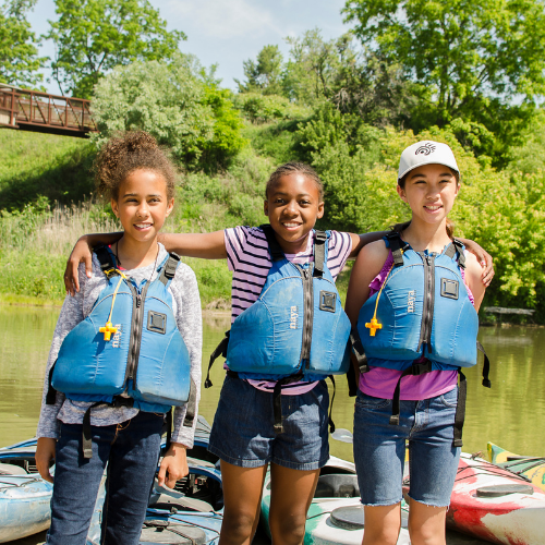 Three children stand facing the camera. They are wearing PFDs and smiling. In the background we see colourful kayaks on the shore of beach, the Rouge River, and green leafy foliage in the distance.