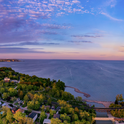 An aerial shot at sunrise looking south over Lake Ontario the southern extent of Rouge National Urban Park. The sky is a mix of light pink, orange, and blues and we can see the beach and surrounding area at the bottom. 